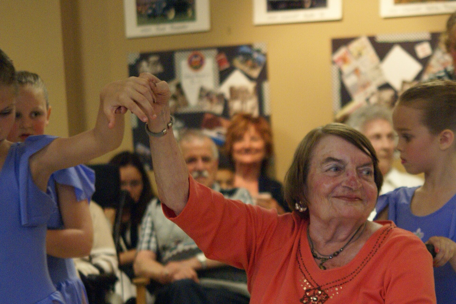 An older woman dancing hand-in-hand with a young girl at a care facility, surrounded by onlookers