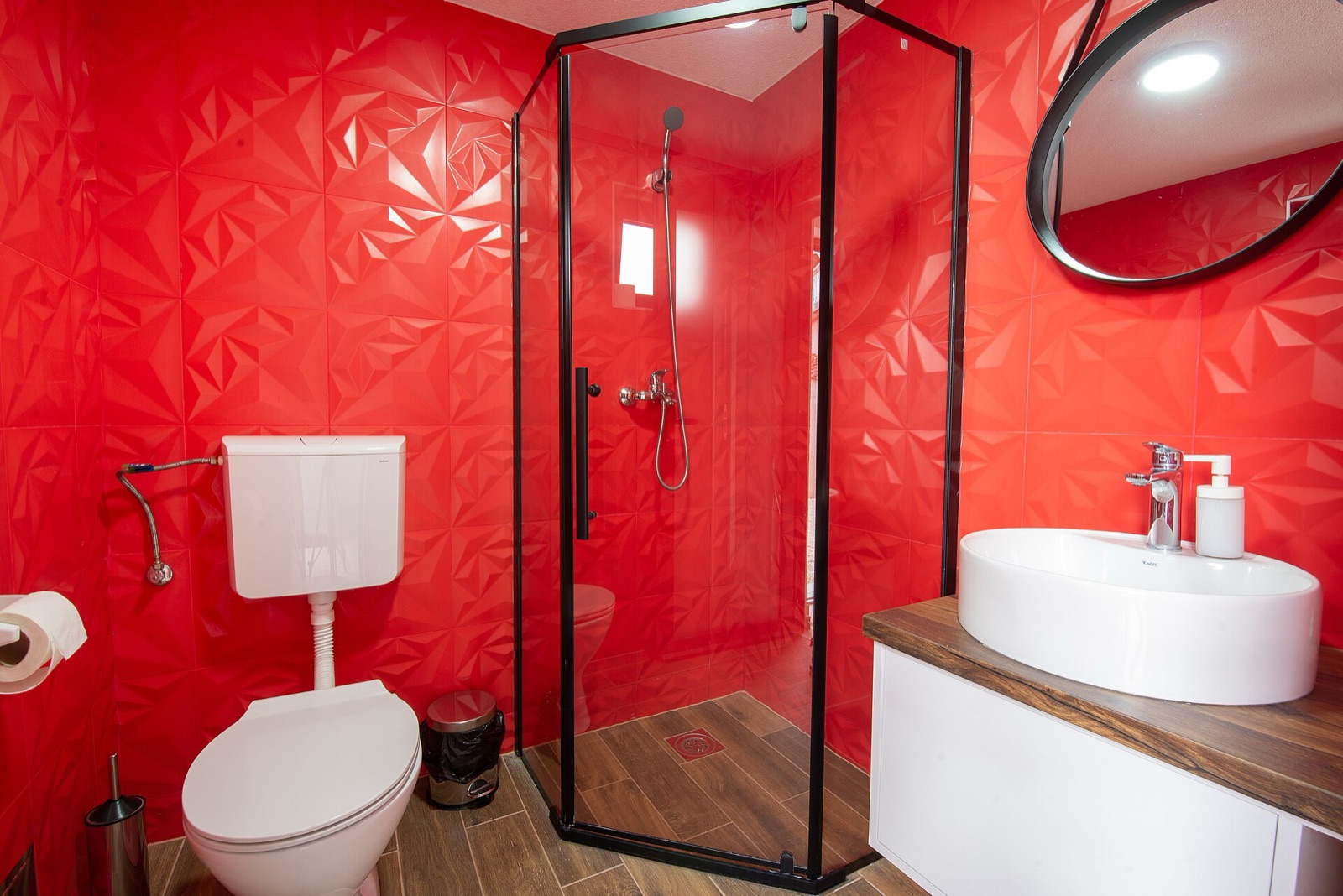 A modern bathroom with saturated red tiled walls, a white toilet, sink, and glass shower