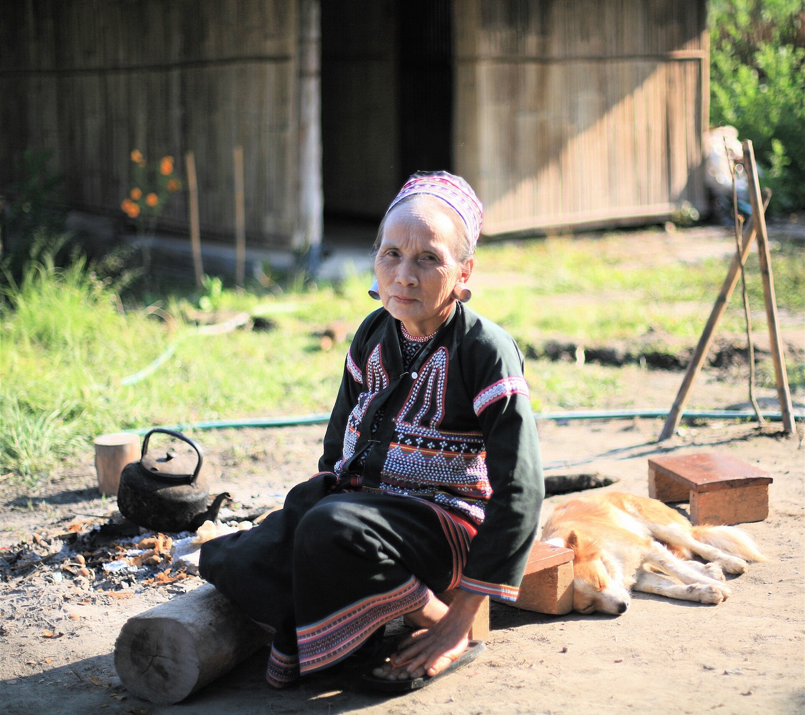 An elderly Lahu woman sitting outside her home in bright sunlight, a kettle on the fire and a dog asleep nearby