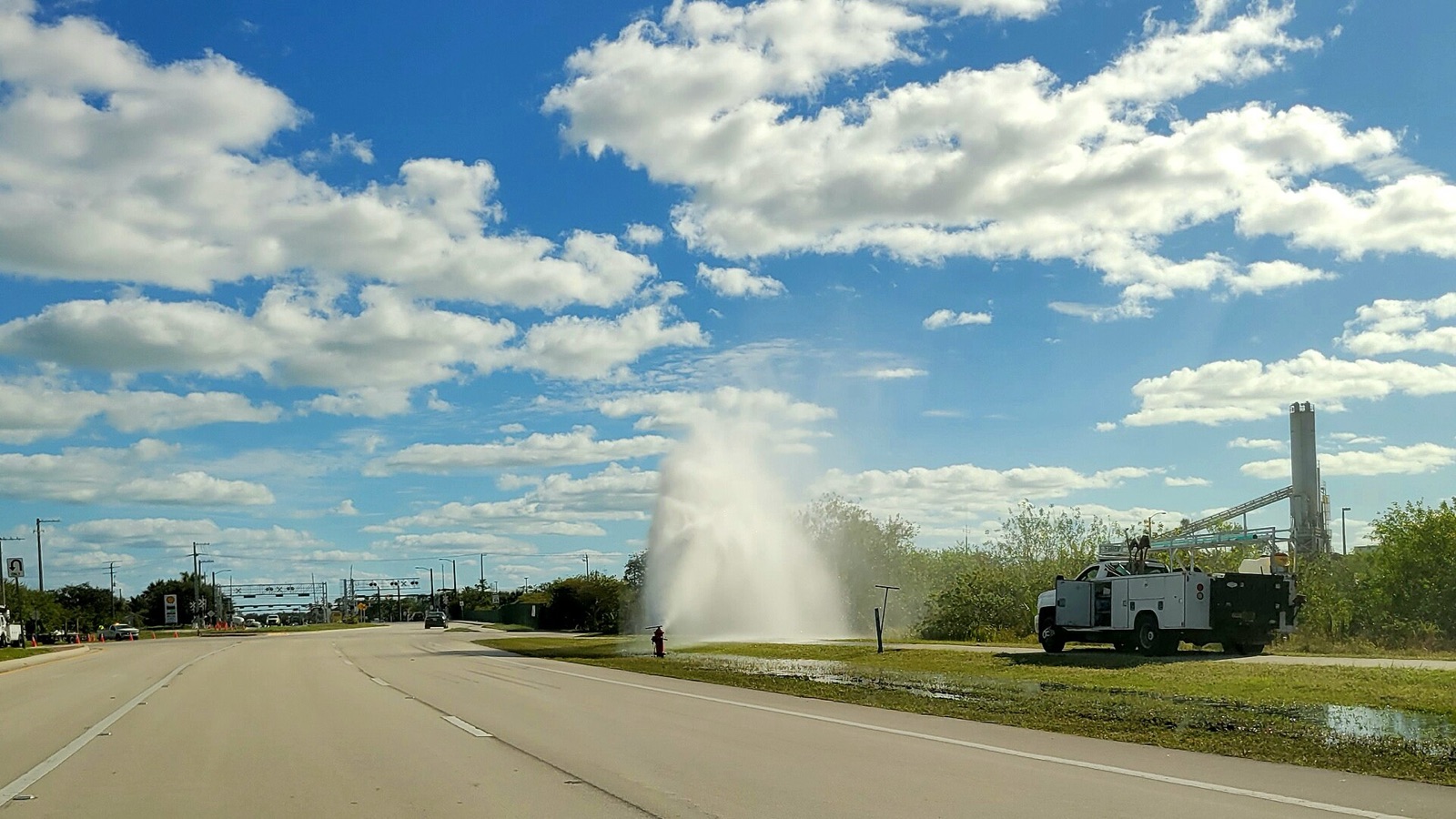 A broken fire hydrant firing a tall plume of water into the air beside a highway, a utility truck parked nearby
