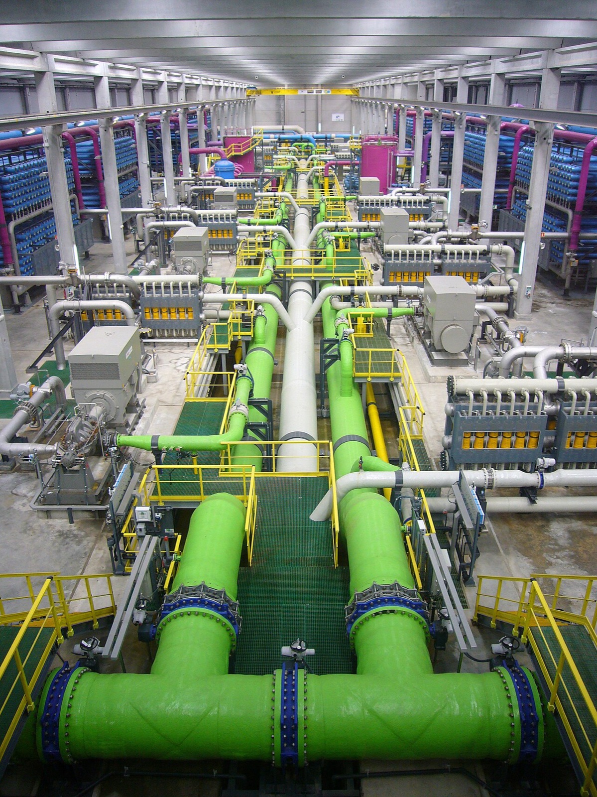 Interior of a reverse osmosis desalination plant: banks of blue pressure vessels flanking huge green feed pipes running down the hall