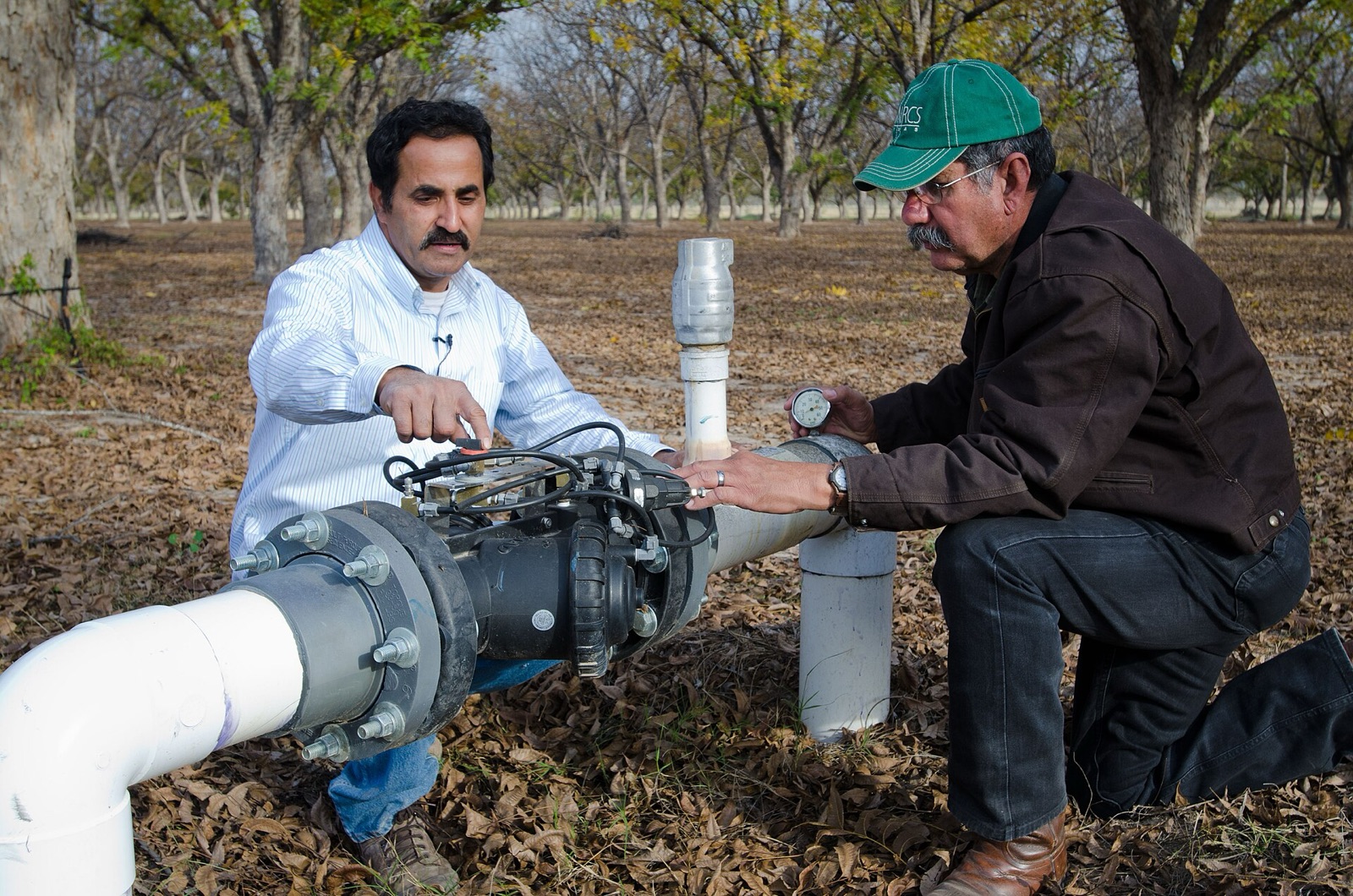 A farmer and a conservation officer kneeling beside a pressure control valve and flow meter in a pecan orchard