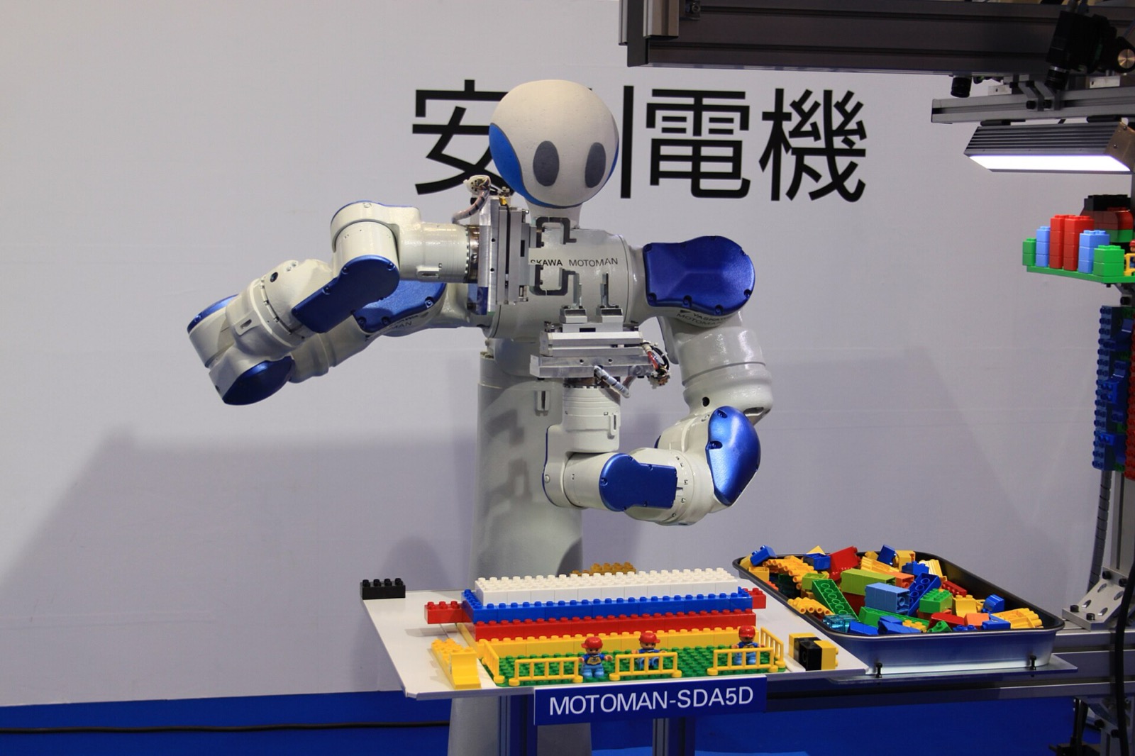 A Yaskawa Motoman dual-arm robot precisely assembling parts beneath the company's logo. Japan's bet is on dexterous, task-built machines, not humanoid showpieces.
